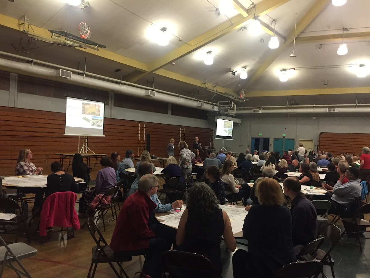 A community meeting in the Cubberley Community Center gym, residents at round tables reviewing site plans